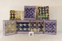 Seven boxes of assorted glass Christmas ornaments arranged in two rows, showing color variety and packaging from Martha Stewart and Sandra brands
