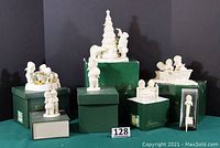 Seven white porcelain Snowbabies figurines and snowglobe all displayed with their original dark green Dept. 56 boxes on a table against a black backdrop.