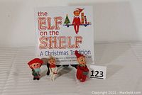Three vintage elf figurines displayed in front of the 'The Elf on the Shelf: A Christmas Tradition' book and an auction number card 123.
