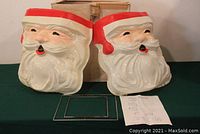 Photo of two blow-mold Santa face pieces placed side by side, metal wire hardware and instructions visible in front, original box in background.