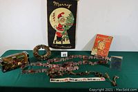 Full lot on green table showing all items including fabric Santa wall hanging, bottle brush wreath, Christmas Carols book, horse and buggy bank, train garlands, and brass bells.