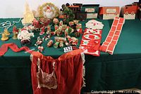 Overview of the entire vintage Christmas decoration collection showing the plastic church, Santa wreath, paper train, ornaments, and apron laid out on a green cloth.