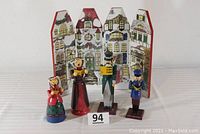 Front view showing 4 wooden carolers and the two-sided village backdrop behind them. Carolers vary in height up to 9 inches, painted in festive colors and holding music sheets.
