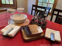 Two napkin holders with napkins, chip and dip serving piece with two white bowls, three large brown plates, four smaller brown plates, flatware in jar caddy, and wooden round tray on red tablecloth at dining table