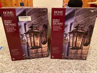 Two boxed Home Decorators Collection Medium Exterior Wall Lanterns with dark ridge bronze finish shown side by side on carpet floor.