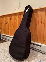 Black soft guitar case standing upright against wooden wall paneling on carpeted floor