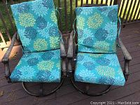 Pair of aluminum outdoor swivel chairs with blue-green floral cushions, placed on wooden deck under sunlight.