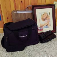 Black Timberland shoulder bag with accessories, wooden lap desk, and green-brown wooden wall frame containing photo of woman and child against pinstriped wood panel wall and carpet floor