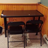 Front view of rectangular desk with two black folding chairs in front, placed against a wood paneled wall and carpeted floor. Child safety straps are visible on desk surface.