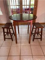 Full view of the round wooden table with two wood stools placed in front of a glass door, showing general shape and size