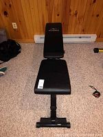 Full view of black adjustable incline workout bench with padded cushions on carpeted floor against wooden wall.