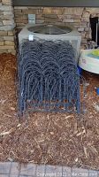 Stacked black metal garden fencing panels placed on wood mulch next to an air conditioning unit