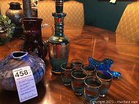 Group of glass items including green Bohemia decanter with stopper, six matching liquor glasses with gold rims and etched designs, cranberry red glass shoe figurine, and blue art glass vase.