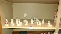 Full view of the eleven white porcelain Snow Babies figurines arranged on a shelf showing various winter and Christmas themed poses.