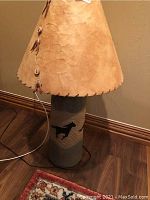 Full view of lamp showing leather shade with button and claw-like decorations, ceramic base, power cord, and lamp height relative to floor molding.
