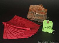 Photo showing eight folded red Beauville napkins with damask pattern next to a piece of petrified wood with auction tag