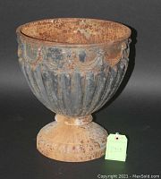 Photo showing a rusted cast iron footed urn with fluted body and decorative garland, set against black background with tag number 2313.
