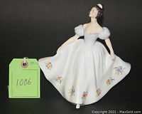 Full view of Royal Doulton Kate figurine standing with arms holding dress, floral decoration on dress hem, white dress with puffy sleeves, 7.75 inches tall.