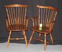Pair of mid-brown stained maple arm chairs by Villas seen frontally against a dark backdrop, showing Windsor-style backs, spindled legs, and armrests.