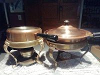 Two copper pans set in two ornate copper stands on table, one stand includes burner, copper lid on large pan visible