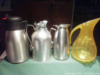 Photo showing all four beverage containers on table with green cloth background, including three stainless steel jugs and one yellow plexiglass pitcher.