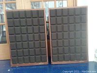 Front view of two rectangular speakers with black fabric grille featuring a square-patterned design.