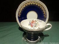 Side angle showing the corset-shaped Aynsley tea cup with cobalt blue exterior, floral interior, and gold trim along rim and base, with matching cobalt and gold saucer in background.