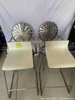 Two modern stainless steel barstools with white contoured seats and decorative circular backs on two stainless steel dining chairs visible in background