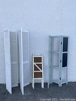 Three white louvered folding doors standing next to a smaller A-frame sign board with white frame and brown panel.