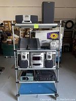 Overview of electronics on shelving including Sony audio system with speakers, Kicker iPhone dock, JVC tuner, vintage slide projector, Philips clock radio, Panasonic boom box