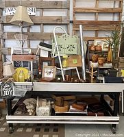 Wide shot showing the large wooden rolling cart with various home decor items on and under it, including framed art, lamps, ceramic cups, glass jugs, wooden trays, and copper salt and pepper shakers.