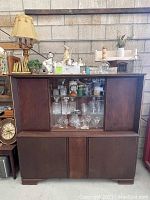 Overall view of closed dark wooden mid century locking cabinet with decorative items on top and visible crystal glassware inside central glass section.
