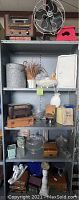 Full view of metal shelving unit containing lot items including vintage radios, Victor fan, breadbox, stool, glassware, ceramics, and books.