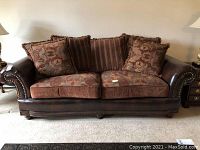 Front view of brown leather couch with patterned brown fabric cushions and throw pillows, showing rolled arms with nailhead trim and wooden legs