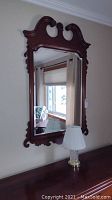 Full view of the solid mahogany framed beveled mirror hung on wall above wooden surface with glass table lamp next to it.