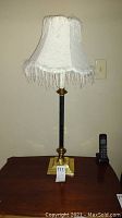 Full view of one lamp showing black and brass base with square bottom and white fabric lampshade with fringe.