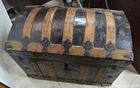 Front and top view of the antique trunk showing curved lid with black metal and wooden bands, decorative metal hardware, and a keyhole.
