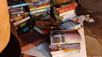 Wide view of multiple stacks of books on a piece of board outside under dim lighting showing various genres and authors.