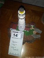 Front angle view of the Boston lighthouse figurine showing the white textured tall tower with yellow light and black railings, attached buildings, grassy green and rocky brown base, on a brown wooden table.