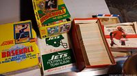 Photo showing multiple boxes including a yellow Score baseball card box, a Fleer baseball card box, and a green New York Jets football card team album along with several storage boxes containing numerous loose sports cards.