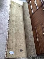 Full view of rolled partially unrolled camel color wool rug with small black designs on a carpeted floor in front of a wood dresser.