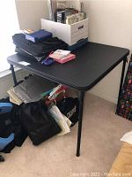 Full view of black folding card table with items placed on top and underneath it, shown in corner of room.