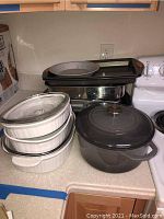 Photo showing three white Corning Ware casserole dishes stacked with glass lids, an enamel-covered dark gray cast iron round pot with glass lid, and Rachel Ray baking and roasting pans on countertop