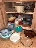 Multiple plastic food storage containers of assorted sizes and lids, Pyrex glass mixing bowls, and various glass dishes displayed in cabinet and on floor