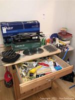 Wide shot of lot showing the Oster electric griddle box, Calphalon griddle box, Deluxe Electric Rotisserie, metal roast holder, assorted cooking utensils, hot pads, and plastic containers on and above the wooden kitchen cart with open drawer of miscellaneous kitchen tools.