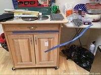 Full view of wood kitchen cart with drawer and cabinet doors, towel racks visible on side, casters at base. Small folding table with plastic top and metal base next to it with miscellaneous kitchen items on top.
