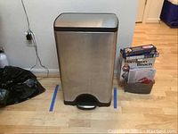 Front view of the stainless steel trash can on wooden floor, showing overall shape and size next to boxes.