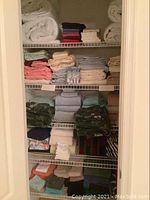 Wide shot of the linen closet shelves showing folded sheets, rolled mattress covers or bed pads, towels, pillow slips, and some patterned fabric pieces on multiple shelves.