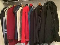 Closet view showing various coats and jackets in different colors hung on hangers, including red, denim blue, tan, black, and cream knit garments.