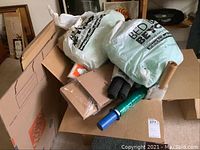 Box filled with cardboard boxes, Bed Bath & Beyond plastic bags, rolls of stretch wrap with blue dispensers, black foam padding rolls and other packing materials.